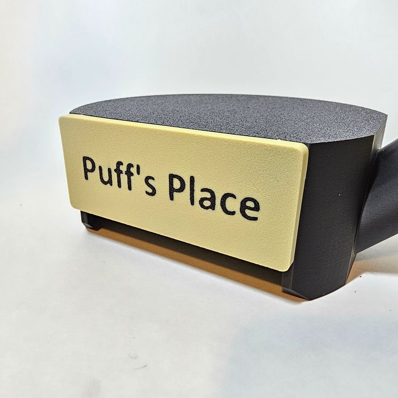 May include: A black and beige object with the text "Puff's Place" on a beige rectangular label. The object has a dark gray, textured top surface and a black base. The background is white.