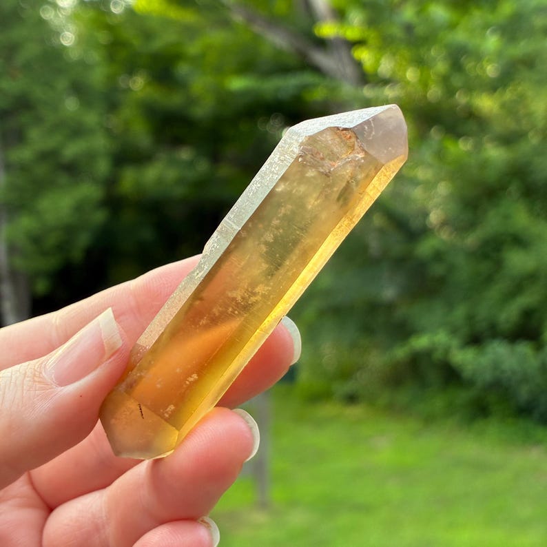 May include: A translucent, elongated, hexagonal crystal point with a vibrant golden-yellow hue. The mineral specimen features a smooth, polished surface with natural internal inclusions and a sharp, faceted termination at the top end.