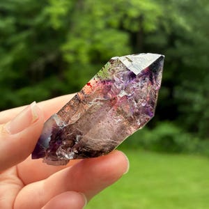 May include: A natural crystal point featuring a blend of deep purple amethyst and smoky brown hues. The translucent mineral displays sharp, angular facets and internal inclusions, with a pointed termination and a jagged, unpolished base structure.