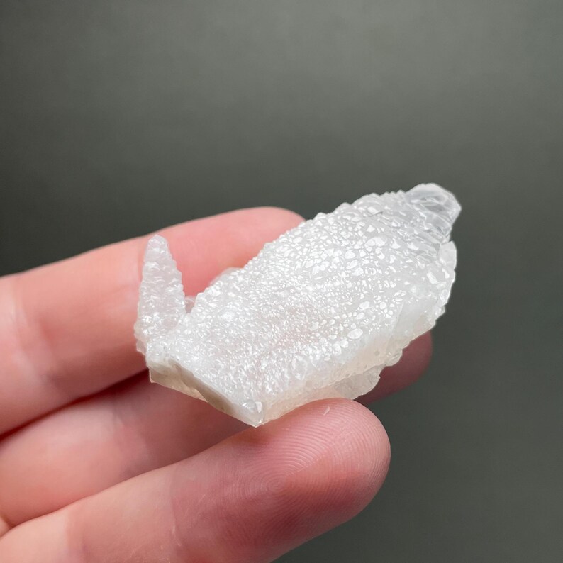 Może przedstawiać: Mały, biały, krystaliczny okaz minerału o teksturowanej powierzchni. Minerał jest trzymany w dłoni na neutralnym tle. Formacja kryształu ma szorstki, nier&oacute;wny wygląd.