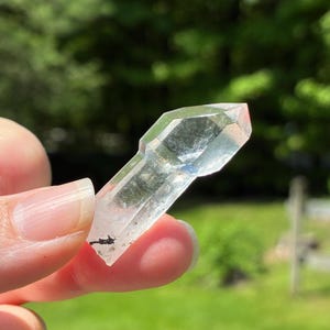 May include: A clear quartz crystal with a faceted, geometric shape. The crystal is held up against a blurred green background. The crystal has a few dark inclusions and is transparent, allowing light to pass through.