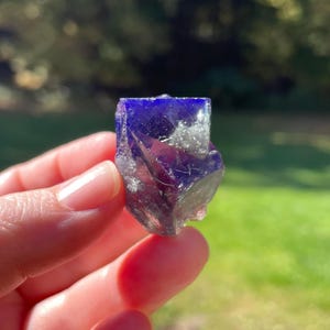 May include: A faceted, deep blue crystal with hints of purple and white. The crystal is held in a hand, with a blurred green background. The crystal's facets catch the light, creating a sparkling effect. The overall theme is natural gemstones.