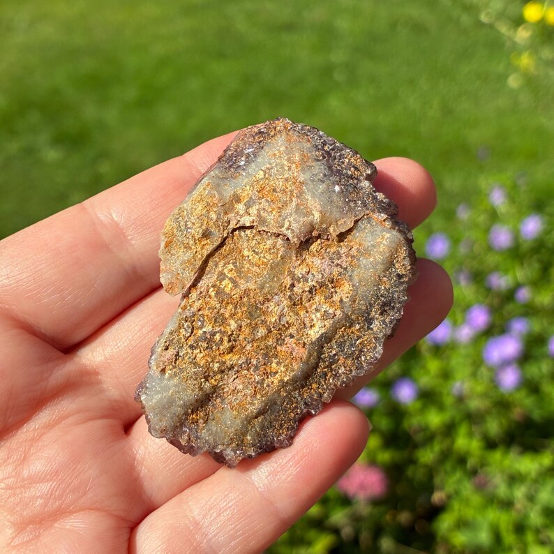May include: A raw, irregular mineral specimen featuring a textured surface with a mix of metallic gold, bronze, and deep purple hues. The stone has a crystalline, jagged edge and a layered appearance with translucent, light-colored mineral deposits.