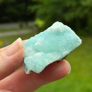 May include: A raw, rectangular-shaped mineral specimen featuring a vibrant, translucent light blue crystalline surface with small, opaque white clusters scattered across the top. The base of the stone appears dark and earthy in texture.