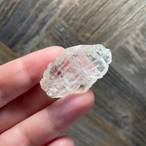 May include: A clear, raw quartz crystal with a rough, textured surface. The crystal is held in a hand, showcasing its natural, irregular shape and translucent quality. The background is a wooden surface.