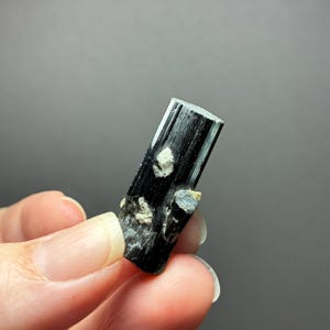Może przedstawiać: Czarny kryształ turmalinu o pionowej, prążkowanej, kolumnowej strukturze. Minerał posiada na powierzchni małe, jasne, nieregularne wtrącenia, co nadaje mu naturalną, surową i niepolerowaną teksturę.
