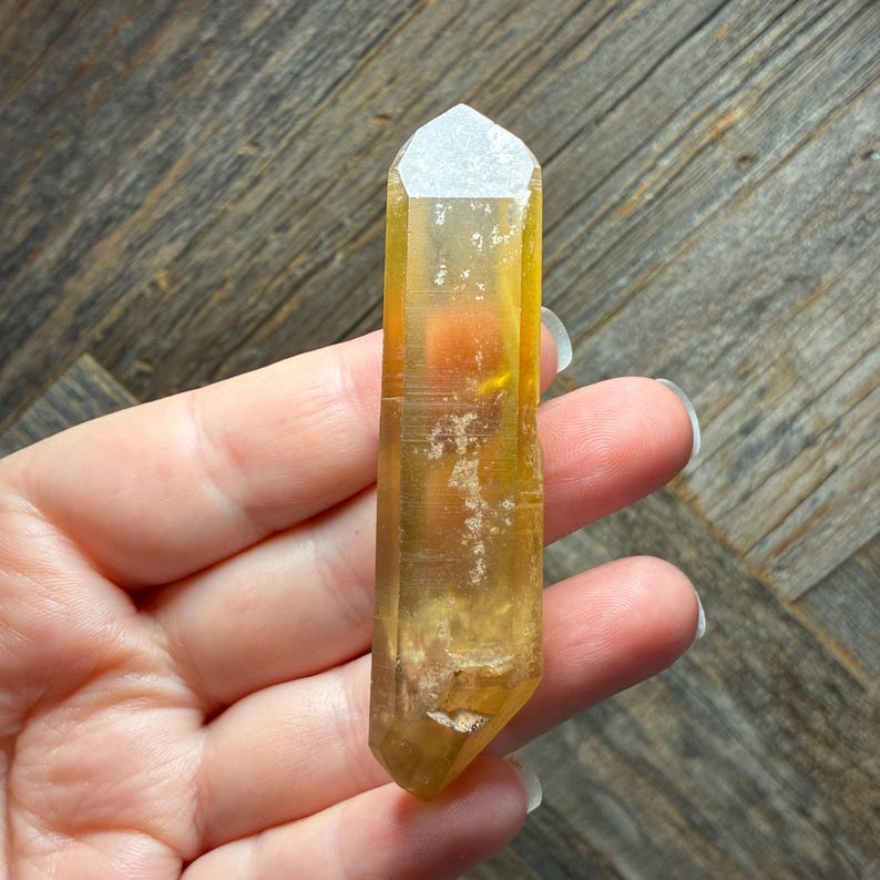 May include: A translucent, elongated citrine crystal point with a hexagonal shape and a pointed termination. The mineral displays a warm, golden-yellow hue with visible internal inclusions, natural fractures, and a smooth, polished surface texture.