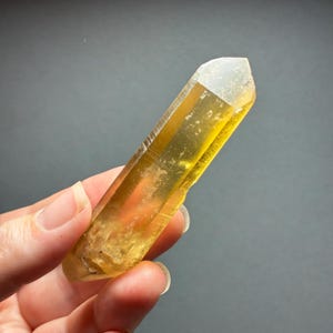 May include: A translucent, elongated citrine crystal point with a hexagonal shape. The stone displays a vibrant golden-yellow hue, featuring natural internal inclusions and a polished, faceted surface that catches the light, showing clear termination.