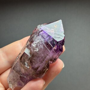 Op de afbeelding: Een ruwe amethist kristalpunt met dieppaarse en doorschijnende violette tinten, natuurlijke interne insluitingen, facetten en een scherpe, puntige uiteinde. Het kristal vertoont een kleurverloop van donkerpaars naar helder transparant.