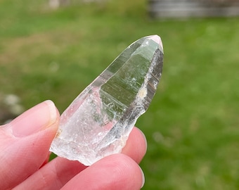 May include: A clear quartz crystal point with a natural, rough base. The crystal is transparent with visible internal structures and a slightly pinkish hue at the tip. The crystal is held against a blurred green background.