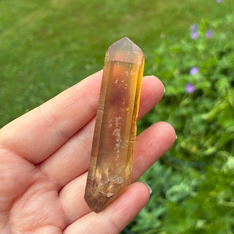 May include: A double-terminated citrine crystal point with a translucent golden-yellow hue. The elongated quartz specimen features natural internal inclusions, visible facets along its length, and a sharp, pointed termination at both ends.