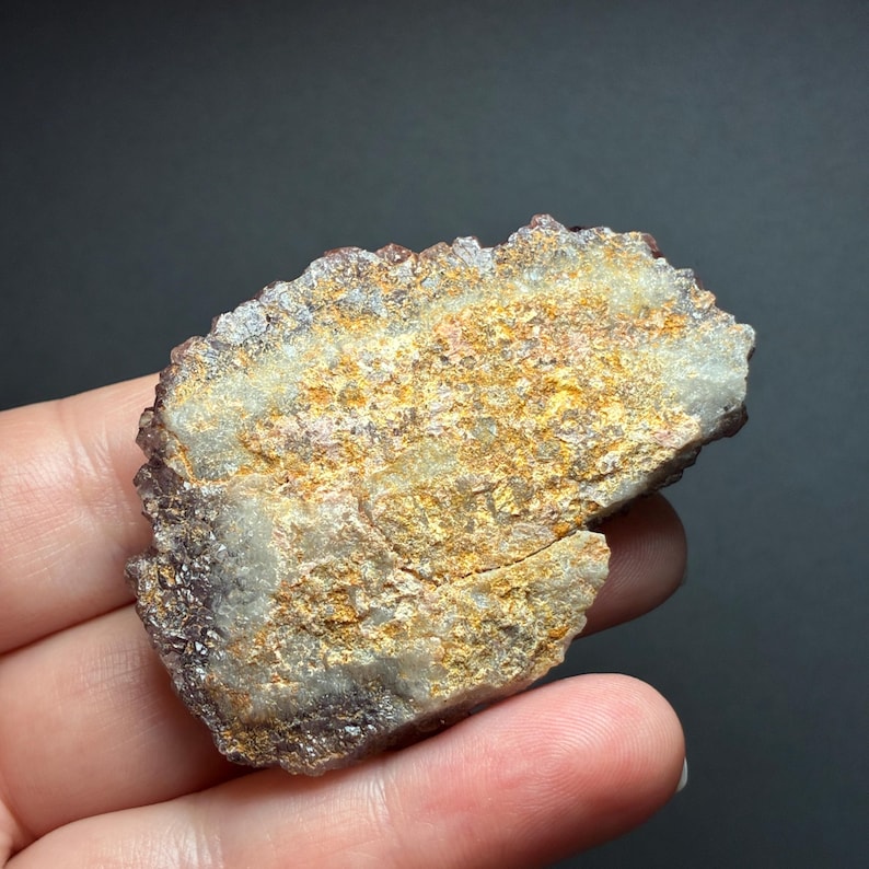 May include: A raw mineral specimen featuring a flat, textured surface with a mix of golden-yellow, tan, and off-white crystalline deposits. The edges of the stone are jagged and dark purple, showcasing a rough, unpolished natural rock formation.