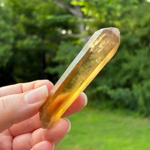 May include: A translucent, elongated citrine crystal point with a hexagonal shape and a warm, golden-yellow hue. The crystal features smooth, polished facets that catch the light, revealing internal clarity and natural variations in its amber color.