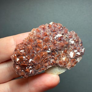 May include: A cluster of raw mineral crystals featuring a deep reddish-brown hue with sparkling, multifaceted surfaces. The specimen displays a dense arrangement of pointed, geometric crystal formations with a light-colored base visible at the bottom.