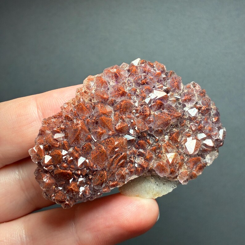 May include: A cluster of raw mineral crystals featuring a deep reddish-brown hue with sparkling, multifaceted surfaces. The specimen displays a dense arrangement of pointed, geometric crystal formations with a light-colored base visible at the bottom.