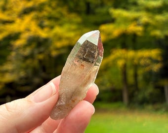 Pode incluir: Uma ponta de cristal de quartzo translúcida e alongada com um tom acinzentado esfumado e inclusões internas visíveis. O cristal apresenta uma terminação pontiaguda naturalmente facetada e uma base texturizada e ligeiramente opaca.