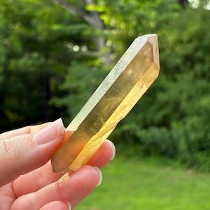 May include: A double-terminated citrine crystal point with a translucent, golden-yellow hue. The elongated, hexagonal mineral specimen features smooth, polished facets that catch the light, displaying natural internal inclusions and clarity.