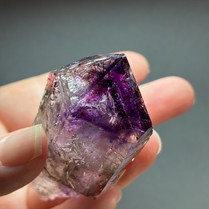 Op de afbeelding: Een doorschijnend amethist kristalexemplaar met dieppaarse kleurzones en interne insluitingen. Het mineraal heeft een geometrische, veelzijdige structuur met scherpe randen en een gepolijst uiterlijk, met natuurlijke variaties in helderheid.