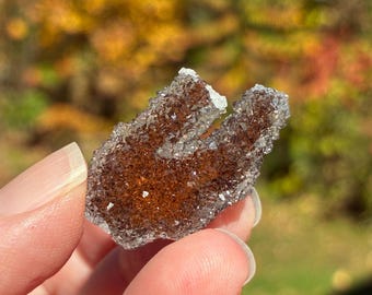 Pode incluir: Um pequeno espécime mineral em forma de U com um centro castanho-âmbar profundo coberto por minúsculos cristais translúcidos e brilhantes. As bordas da formação são definidas por uma textura cristalina irregular e serrilhada.