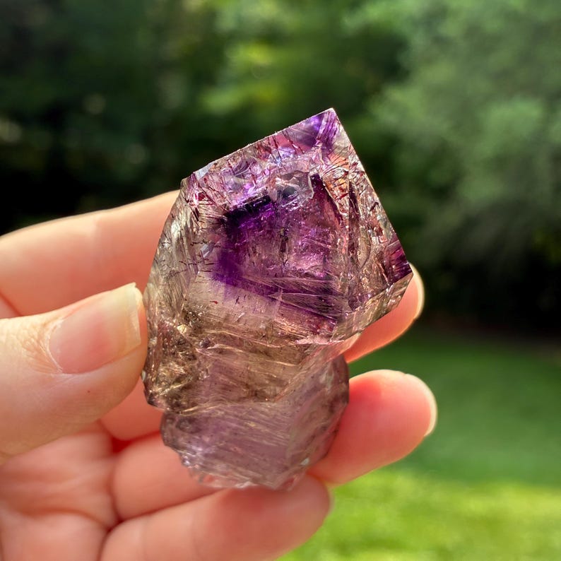 Op de afbeelding: Een ruwe amethist kristalpunt met dieppaarse tinten aan de top, die overgaan in een rokerige, doorschijnende basis. Het mineraal vertoont natuurlijke interne breuken, scherpe geometrische facetten en een glanzende, semi-transparante textuur.