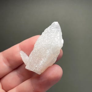 Może przedstawiać: Surowy, naturalny biały klaster kryształ&oacute;w o teksturowanej powierzchni. Kryształ ma spiczastą końc&oacute;wkę i mniejszą, wt&oacute;rną formację kryształ&oacute;w rozciągającą się z boku. Tło jest jednolite, stonowane szare.