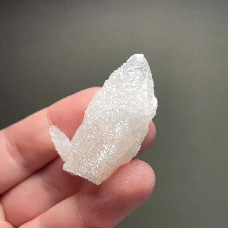 Może przedstawiać: Surowy, naturalny biały klaster kryształ&oacute;w o teksturowanej powierzchni. Kryształ ma spiczastą końc&oacute;wkę i mniejszą, wt&oacute;rną formację kryształ&oacute;w rozciągającą się z boku. Tło jest jednolite, stonowane szare.