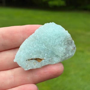 May include: A raw, light blue mineral specimen with a crystalline, textured surface. The stone features a small, dark brown inclusion embedded within the lower section of its translucent, jagged structure, showcasing natural mineral formations.