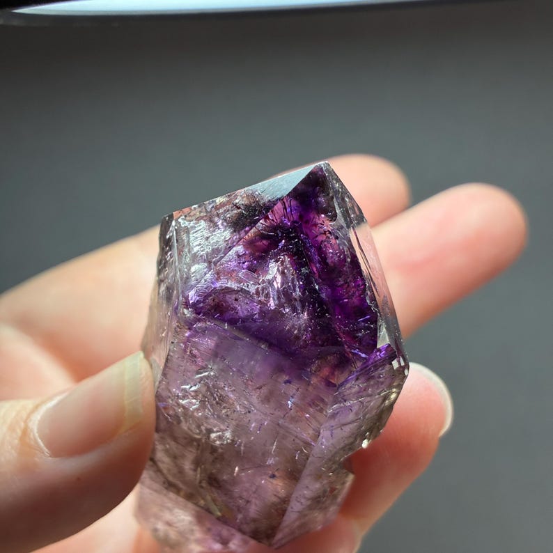 Op de afbeelding: Een doorschijnend, puntig kristalexemplaar met dieppaarse fantomen en insluitingen binnen zijn heldere, gefacetteerde structuur. Het mineraal vertoont scherpe geometrische randen en een duidelijk kleurverloop, overgaand van een donkerpaarse kern naar een lichtere, transparante basis met zichtbare interne breuken.