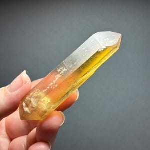 May include: A double-terminated crystal point with a translucent yellow hue. The elongated mineral specimen features a faceted hexagonal structure with visible internal inclusions and natural surface textures, transitioning from a clear tip to a deep golden base.