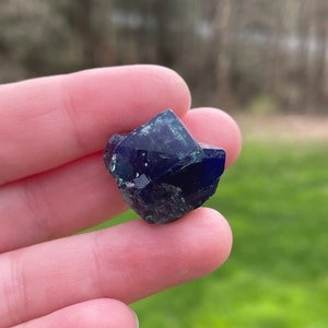 May include: A close-up of a dark blue crystal with sharp, geometric facets. The crystal has a deep, saturated color with hints of green in some areas. The background is blurred, suggesting an outdoor setting.
