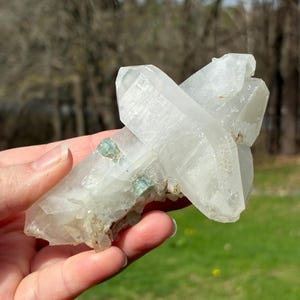 May include: A clear quartz crystal cluster with multiple points, held in a hand. The crystal has a translucent appearance with some areas of green inclusions. The background is blurred, showing a natural outdoor setting.