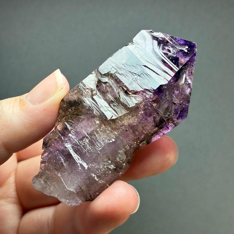 Op de afbeelding: Een ruwe amethist kristalpunt met een natuurlijke zeshoekige vorm en dieppaarse, doorschijnende violette tinten. Het oppervlak vertoont grillige, ongepolijste facetten en interne insluitingen die een duidelijke structuur tonen.