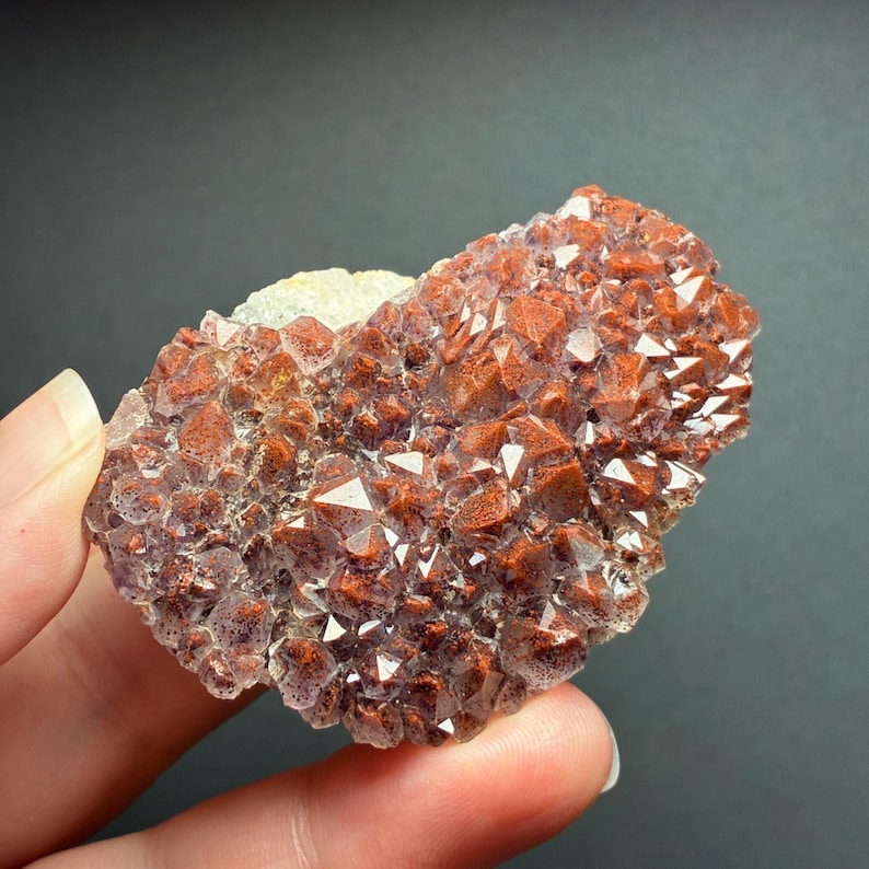 May include: A cluster of raw mineral crystals featuring a dense arrangement of pointed, reddish-brown and translucent purple points. The specimen has a rough, textured surface with a light-colored base visible on one side of the crystalline formation.