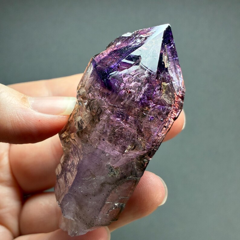 Op de afbeelding: Een ruwe amethist kristalpunt met dieppaarse en doorschijnende violette tinten. Het natuurlijke mineraalexemplaar vertoont een scherpe, gefacetteerde punt aan de bovenkant met complexe interne breuken, grillige randen en donkere insluitingen.