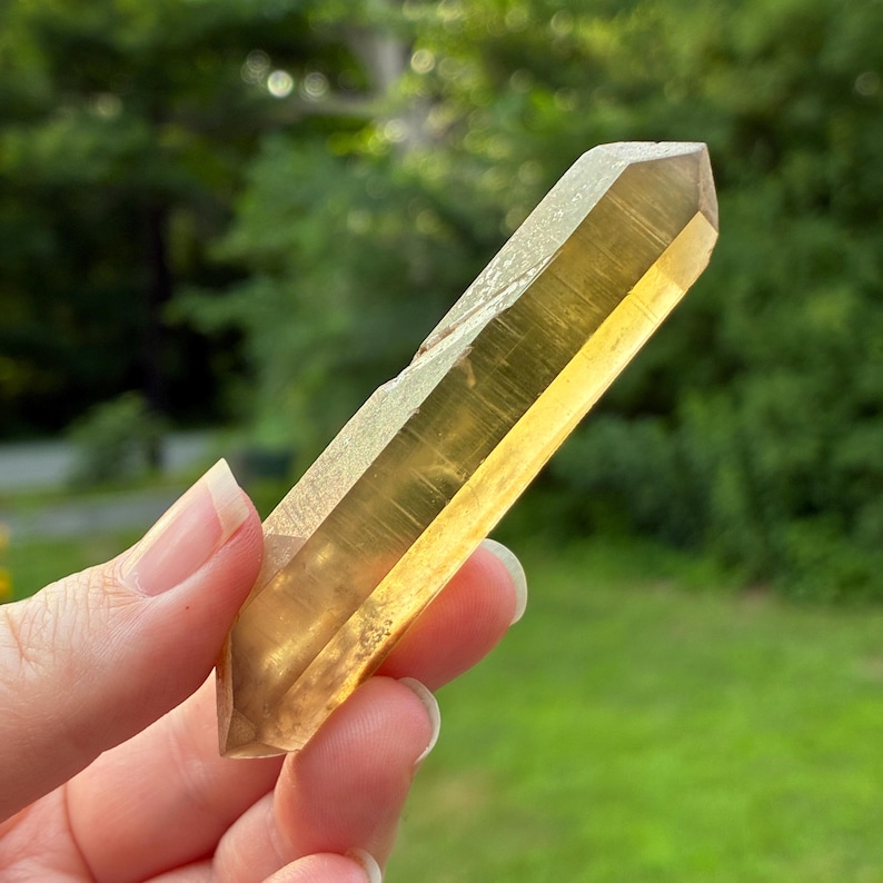 May include: A double-terminated citrine crystal point with a translucent, golden-yellow hue. The quartz specimen features a hexagonal prismatic shape with naturally faceted ends, displaying internal clarity and subtle linear striations along its length.