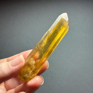 May include: A translucent, elongated, hexagonal crystal point with a vibrant golden-yellow hue. The mineral specimen features a sharp, faceted termination at one end and a textured, cloudy base, displaying natural internal inclusions and clarity.