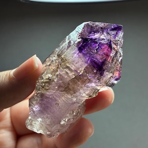 Op de afbeelding: Een doorschijnende amethist kristalpunt met een dieppaars fantoominterieur, natuurlijke grillige randen en een gefacetteerd oppervlak. Het kristal vertoont een verloop van lichtviolette tot donkerpaarse tinten met zichtbare insluitingen.