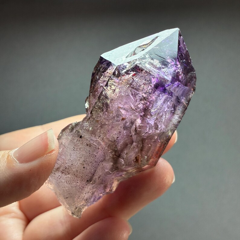 Op de afbeelding: Een ruwe amethist kristalpunt met een doorschijnende paarse tint, diepviolette insluitingen en een scherpe, gefacetteerde top. Het mineraal vertoont natuurlijke geometrische facetten, interne breuklijnen en een verloop van tinten.