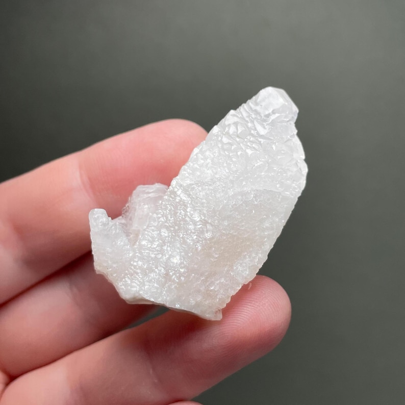 Może przedstawiać: Zbliżenie białego, krystalicznego okazu mineralnego. Minerał ma szorstką, teksturowaną powierzchnię i nieregularny kształt. Kolor jest blady, p&oacute;łprzezroczysty biały, z niekt&oacute;rymi obszarami wydającymi się bardziej nieprzezroczystymi. Okaz jest trzymany w dłoni na szarym tle.