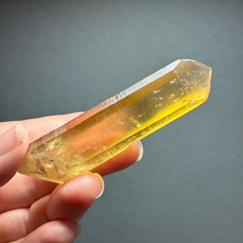 May include: A translucent, elongated citrine crystal point with a hexagonal shape and a pointed termination. The mineral displays a vibrant golden-yellow hue, featuring natural internal inclusions and smooth, reflective facets along its length.