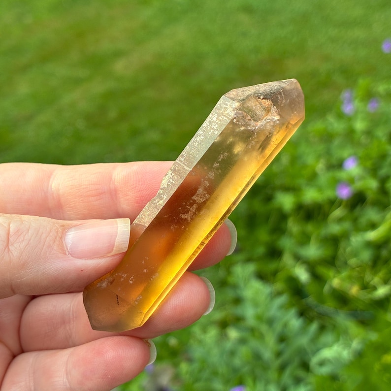 May include: A translucent, elongated citrine crystal point with a warm, golden-yellow hue. The natural mineral specimen features a faceted, hexagonal structure with a pointed termination and visible internal inclusions within the amber-toned body.
