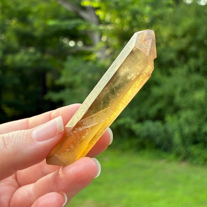 May include: A translucent, golden-yellow crystal point with a hexagonal shape and a pointed termination. The mineral exhibits internal inclusions and a smooth, polished surface, reflecting light through its amber-toned, elongated structure.
