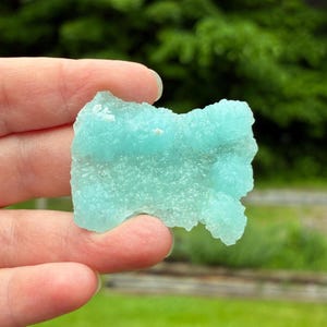 May include: A raw, translucent light blue mineral specimen with a crystalline, textured surface. The stone features an irregular, jagged shape with small, clustered formations across its face, displaying a vibrant, cool-toned turquoise hue.