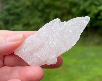 May include: A clear, crystalline quartz cluster with a rough, textured surface. The quartz is held against a blurred green background, highlighting its natural, irregular shape. The stone appears translucent, catching the light.
