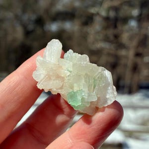 May include: A cluster of translucent, crystalline minerals, primarily white with hints of pale green. The crystals have a rough, textured appearance, and are held in a person's hand against a blurred outdoor background.
