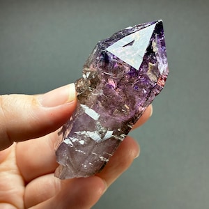 Op de afbeelding: Een natuurlijke amethist kristalpunt met dieppaarse en doorschijnende violette tinten. De steen heeft een scherpe, gefacetteerde punt met zichtbare interne insluitingen, natuurlijke breuken en een gepolijste, reflecterende oppervlaktestructuur.