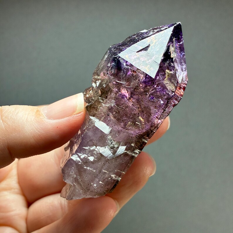 Op de afbeelding: Een natuurlijke amethist kristalpunt met dieppaarse en doorschijnende violette tinten. De steen heeft een scherpe, gefacetteerde punt met zichtbare interne insluitingen, natuurlijke breuken en een gepolijste, reflecterende oppervlaktestructuur.