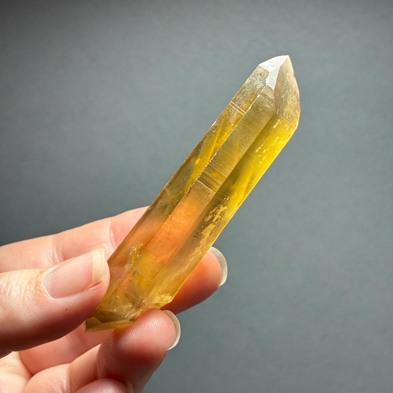 May include: A translucent, elongated citrine crystal point with a vibrant golden-yellow hue. The mineral specimen features a naturally faceted, hexagonal structure with a sharp, pointed termination and visible internal inclusions and fractures.