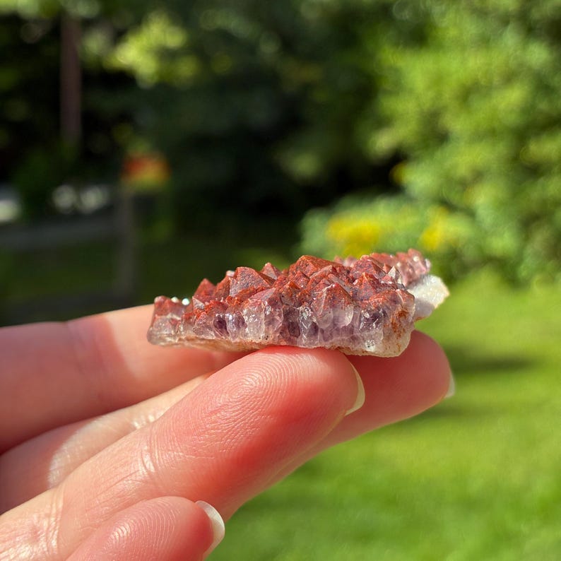 May include: A cluster of small, pointed crystals featuring a deep purple base that transitions into a reddish-brown, iron-stained coating on the tips. The mineral specimen has a jagged, uneven surface with multiple crystalline points.