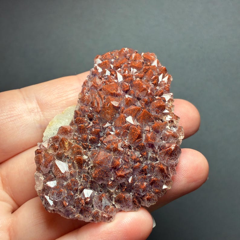 May include: A cluster of small, pointed crystals with a deep reddish-brown hue and translucent edges. The mineral specimen features a textured, jagged surface with numerous sharp, multifaceted points that catch the light, displaying a mix of dark rust and clear crystalline structures.